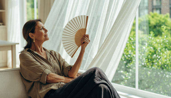 A calm woman in her fifties cooling off in a bright, airy living room by a sunlit window on a warm summer day
