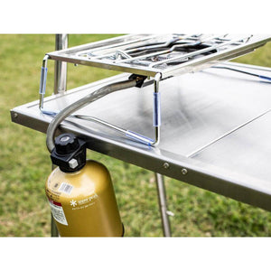Snow Peak camping stove attached to a metal table with a fuel canister hanging below