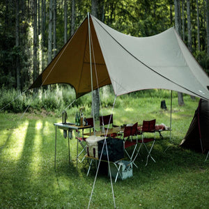 Camping setup with a large tarp, folding table, chairs, and cooking equipment in a forest clearing