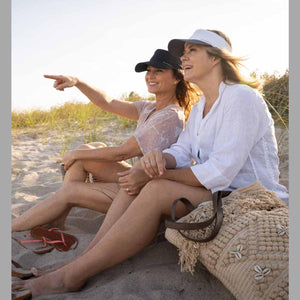 Two women wearing Mission visors sitting on a sandy beach