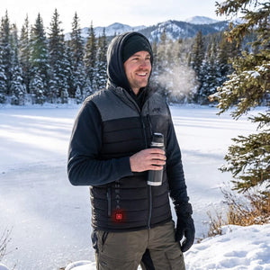 Smiling man in a black and grey heated vest, showing "HEAAT A" and a glowing red power button, holds a thermos in a bright, snowy winter landscape.