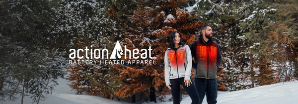 Two people wear ActionHeat battery-heated vests with visible red glowing elements, standing in a snowy evergreen forest with the 