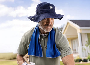 A man in a "MISSION" sun hat and blue cooling towel works outdoors, holding a wooden tool handle, with a house in the background.