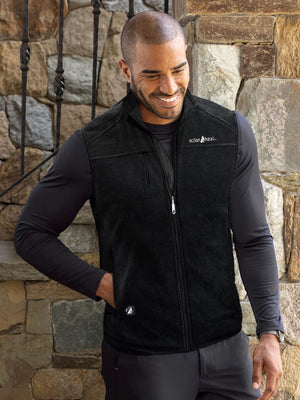 A smiling man in a black long-sleeved shirt and black fleece vest with "action heat" logo, hand in pocket, in front of a stone wall.
