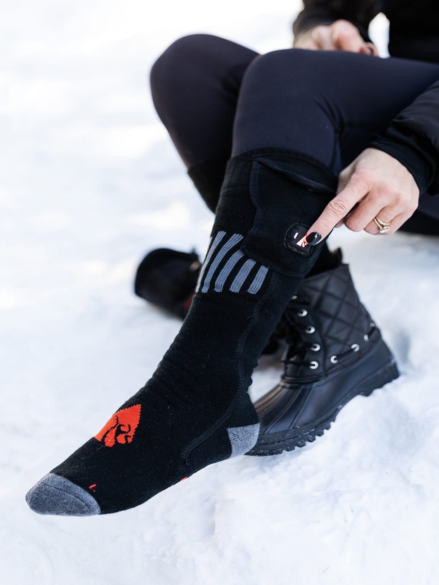 A person sits in snow, pressing the glowing flame button on a black heated sock with gray stripes and a red logo, worn with black quilted boots.