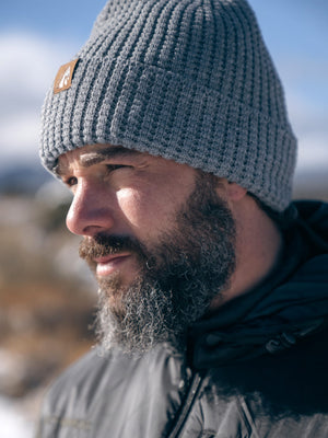 Close-up profile of a bearded man wearing a gray beanie with a brown mountain logo patch, looking left against a bright, blurred blue sky.