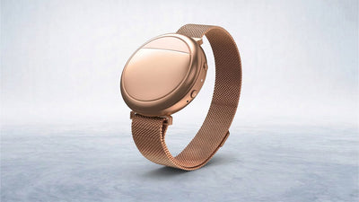A sleek rose gold smartwatch with a round face and metallic mesh strap rests tilted on a light blue-grey surface.