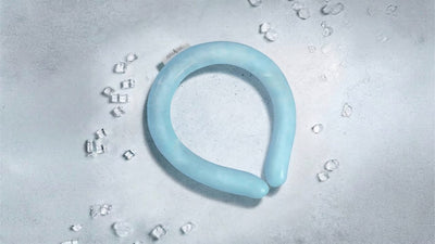 Light blue U-shaped cooling neck collar on a grey textured surface, surrounded by scattered clear ice cubes. A small tag reads 'alpha Cool'.