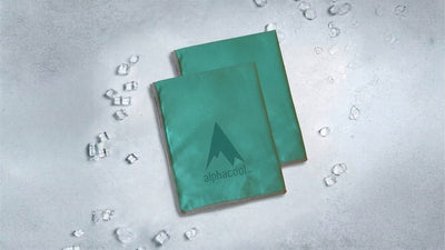 Two teal Alphacool cooling towels, with a mountain logo on one, rest on a grey surface scattered with ice cubes.