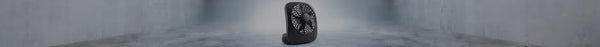 A small, dark grey portable fan with a textured grille stands centered on a seamless light grey concrete floor and wall.