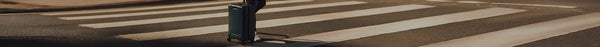 A dark blue suitcase with wheels stands on a zebra crossing with white stripes on asphalt under bright sunlight, casting a shadow.