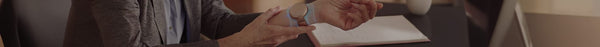 A person in a brown sweater adjusts a tan circular device on a light blue band worn on their left forearm, above an open notebook on a dark desk.