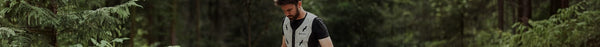A bearded man wearing a light grey vest and black t-shirt looks down, standing in a dense green forest.