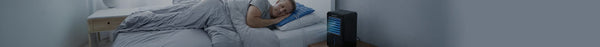 A man sleeps soundly in a dim bedroom, covered by a grey duvet, with a blue cooling pillow and a black portable cooler emitting blue light nearby.