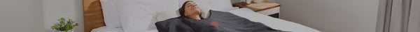 A woman with closed eyes lies peacefully on her back in a bed with white sheets, covered by a dark blanket featuring a red flame logo.