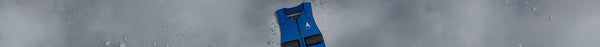 A bright blue flotation vest with a white mountain logo and black straps stands vertically against a wet, light grey surface with many water droplets.