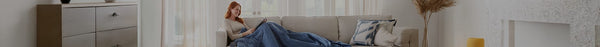 A smiling woman with red hair relaxes under a blue blanket on a beige couch in a naturally lit living room with a wooden dresser and patterned wall.