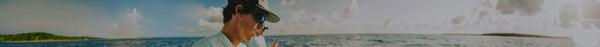 A man in a hat and blue sunglasses fishes from a boat on the water, with distant green islands under a partly cloudy sky.