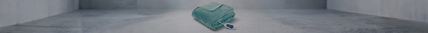 A neatly folded, plush light blue electric blanket with a white and blue controller rests on a grey concrete floor in a minimalist room.