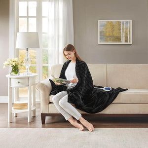 Woman sitting on a sofa reading a magazine while wrapped in a black Beautyrest electric throw blanket