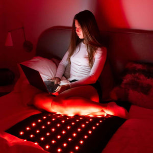 Woman sitting on a bed using a laptop with a red light therapy mat on her lap