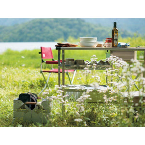Snow Peak metal camping table with shelves and cookware set up in a grassy field by a lake