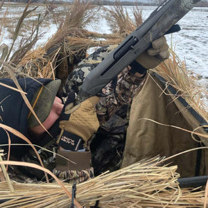 Hunter in camouflage gear wearing Glacier Glove waterproof gloves holding a shotgun in a grass blind
