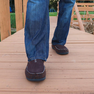 Person wearing brown Acorn slippers with blue jeans on a wooden deck
