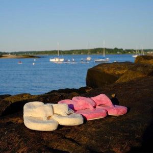 Acorn white and pink plush flip flops on a rock by the water