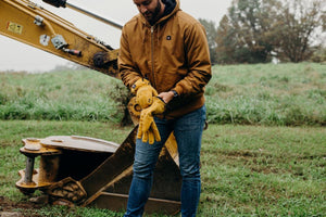 Man wearing ActionHeat brown work jacket putting on yellow leather work gloves by construction equipment