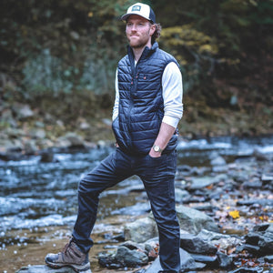 Man wearing a black ActionHeat heated vest over a white long sleeve shirt and jeans standing by a stream