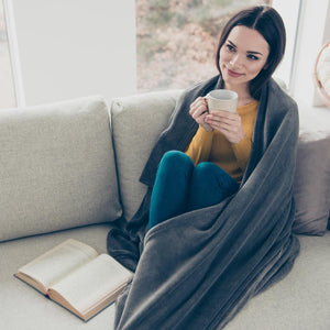 Woman sitting on a sofa wrapped in an ActionHeat grey blanket holding a mug with an open book nearby