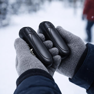 Person wearing grey gloves holding two black ActionHeat hand warmers in a snowy environment