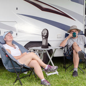 AlphaCool portable fan on a table between two people sitting in camping chairs outside a recreational vehicle