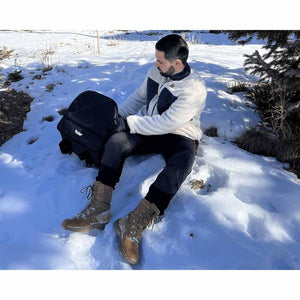 Man sitting in snow holding a black AO Coolers backpack