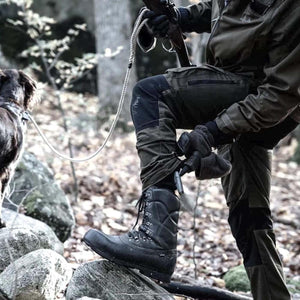 Arctic Volt hunter adjusting black lace up boot in forest with dog on leash