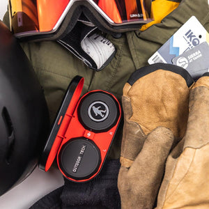 Outdoor Tech wireless audio chips in a charging case surrounded by ski gear