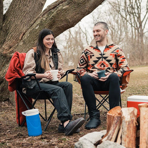 Couple sitting in camping chairs by a fire pit with Coleman cooler and mugs