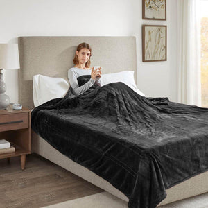 Person sitting in bed under a dark grey Beautyrest heated throw blanket holding a mug
