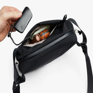 Hand holding a Bellroy key cover over an open black crossbody bag containing small personal items