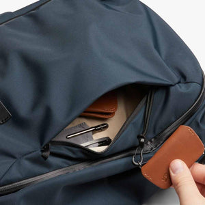 Hand opening a zippered pocket on a blue Bellroy backpack containing a wallet and pens