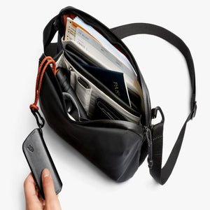 Open Bellroy sling bag containing travel documents and a passport with a hand holding a leather key cover