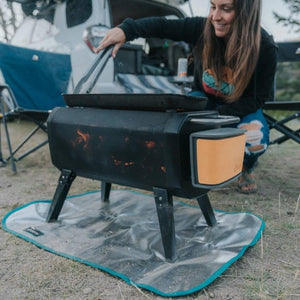 Person using a BioLite FirePit with a griddle top on a heat shield mat at a campsite
