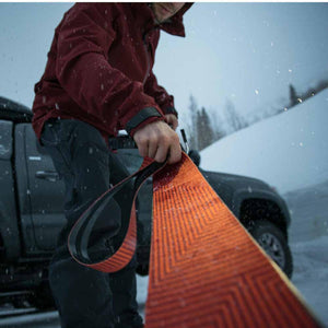 Person in red jacket holding orange climbing skin for skis next to a truck in the snow