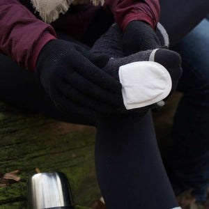 Person wearing black gloves applying a white Hothands toe warmer to a grey sock