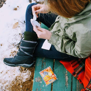 Person opening a Hothands toe warmer packet while sitting on a bench in the snow