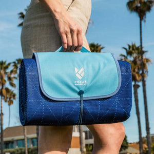 Person carrying a folded blue CGEAR sand-free mat with a light blue flap and logo