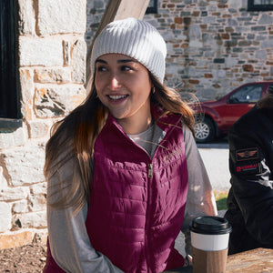 Woman wearing a white beanie and a maroon Venture Heat vest sitting at an outdoor table with a coffee cup