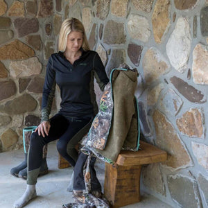 Woman wearing black and olive base layer set sitting on wooden bench with camouflage jacket