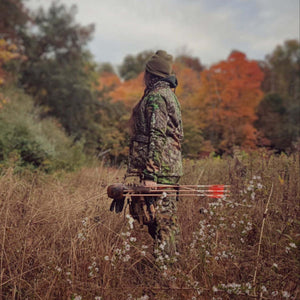 Person wearing camouflage hunting gear standing in a field holding a quiver of arrows